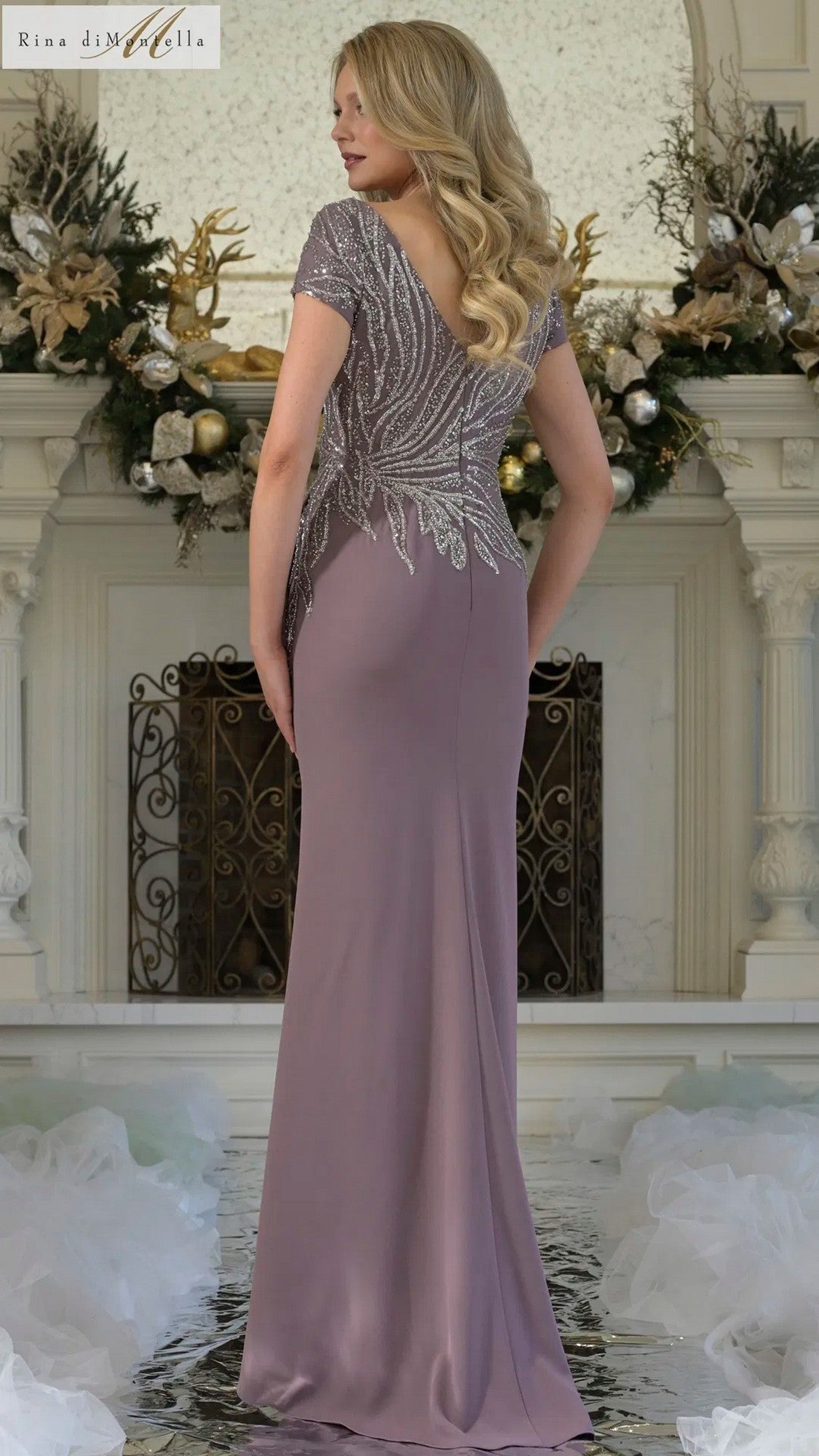 A woman wearing the Long Formal Dress: Rina Di Montella RD3323, featuring a beaded bodice, stands indoors facing away from the camera before an ornate white fireplace decorated with greenery and ornaments.